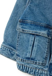 Tela de mezclilla, color azul, presenta un amplio bolsillo con solapa y puño elástico. La textura está ligeramente desgastada con costuras en contraste.