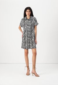 Μαύρο και άσπρο φόρεμα με σχέδιο, κοντά μανίκια, σφιχτή μέση και γιακά. Συνδυασμένο με καφέ πέδιλα με λουράκια, τοποθετημένο σε ουδέτερο φόντο.