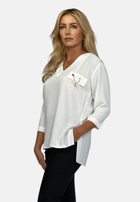 Biała bluzka z dekoltem w serek i rękawami o długości trzech czwartych, z kieszenią ozdobioną łańcuszkiem. Noszona z czarnymi spodniami, materiał wydaje się miękki i teksturowany.