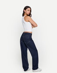 Jean large bleu foncé en denim, avec une texture douce, une taille mi-haute et des coutures contrastées. Stylisé avec un haut court blanc.
