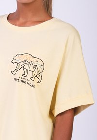 Gelbes Baumwoll-T-Shirt mit lockerer Passform. Es zeigt ein Bärenmotiv mit Bergen, Sonne und Bäumen, begleitet von dem Text "GEBOREN UM MEHR ZU ERKUNDEN."