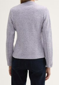 Kvinna klädd i en åtsittande ljusgrå långärmad jacka och mörkblå jeans, sedd bakifrån mot en enkel bakgrund.