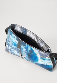 Niebieska torba z nadrukiem i zamkiem błyskawicznym u góry, prezentująca mieszankę wzorów graficznych i tekstur. Czarny pasek oraz smukły, kompaktowy kształt.