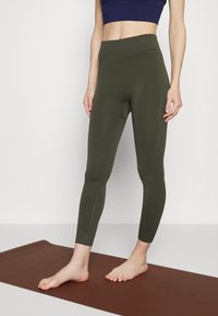 Osoba ubrana w oliwkowe legginsy i granatowy crop top, stojąca boso na brązowej macie do jogi na tle białym.