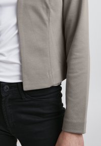 Ljusgrå kort jacka med en slät yta, tillsammans med åtsittande svarta jeans med knappstängning och diskreta fickdetaljer.