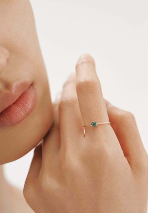 Anillo de oro con una pequeña gema verde, banda delgada, diseño minimalista, usado en un dedo cerca de la cara de una persona, con tonos suaves de piel visibles.