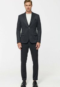 Férfi sötétszürke, karcsúsított öltönyt visel fehér inggel és fekete elegáns cipővel, egyszerű fehér háttér előtt áll.