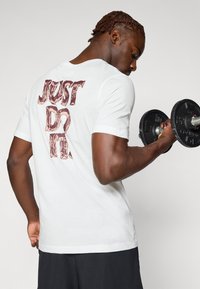 Hvid t-shirt med korte ærmer og rødt "JUST DO IT" grafik på ryggen, blødt stof, løs pasform, brugt mens der løftes vægte.