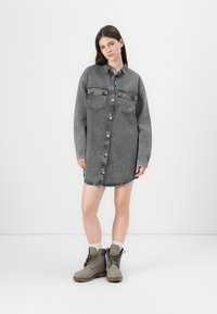 Grå denim skjortekjole med lange ermer, knappefront, to brystlommer og en frynset nederkant. Kombinert med grå støvler og hvite sokker.