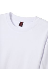 Camiseta de algodón blanca con cuello redondo, que presenta una etiqueta negra cosida en el escote. Los detalles de las costuras son visibles en el cuello y las mangas.