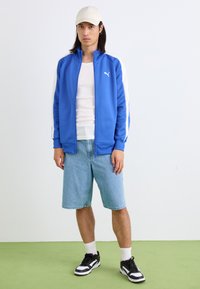 Jachetă sport albastră cu fermoar, cu accente albe, asortată cu pantaloni scurți din denim deschis și adidași alb-negru. Modelul poartă o șapcă bej.