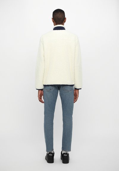 Cream fleece jacket with a navy collar, relaxed fit, and textured surface paired with light blue jeans and black shoes.