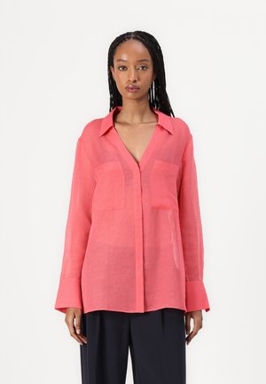Blouse corail transparente avec un col en V, des manches longues, deux poches poitrine et des poignets boutonnés, associée à un pantalon large sombre.