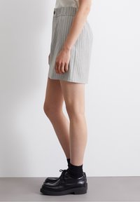 Pantalones cortos gris a rayas con frente plano y cierre de botón, combinados con zapatos negros de cordones y calcetines negros. Vista de perfil lateral.