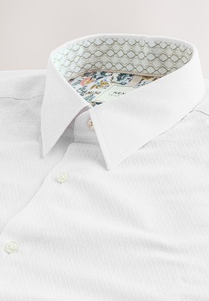 Camicia bianca con superficie strutturata, colletto a punta e interno decorativo con motivo floreale. Due chiusure con bottoni visibili.