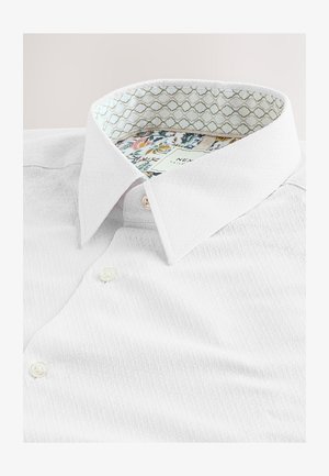 Chemise blanche en tissu texturé, avec col pointu et doublure intérieure décorative à motifs floraux. Fermeture avec deux boutons visibles.