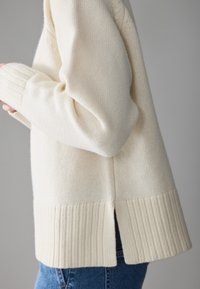 Pull en maille couleur crème avec ourlet et poignets côtelés, présentant des fentes sur les côtés. Texture douce et coupe décontractée, porté avec un jean bleu.