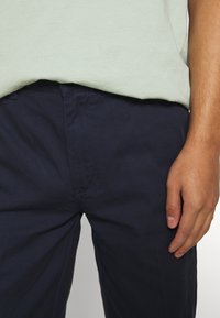 Mörkblå bomullsbyxor med rak bendesign, som har synlig sömnad och en jämn textur, i kombination med en ljusgrön skjorta.