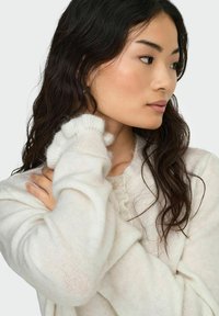 Maglione bianco lavorato a maglia con scollo arricciato, dettagli con bottoni e maniche leggermente arrotolate, dal finish morbido e testurizzato.