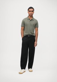 Polo vert olive avec un petit logo, associé à un pantalon noir à jambes larges et des baskets blanches à semelles beiges. Design simple et épuré.