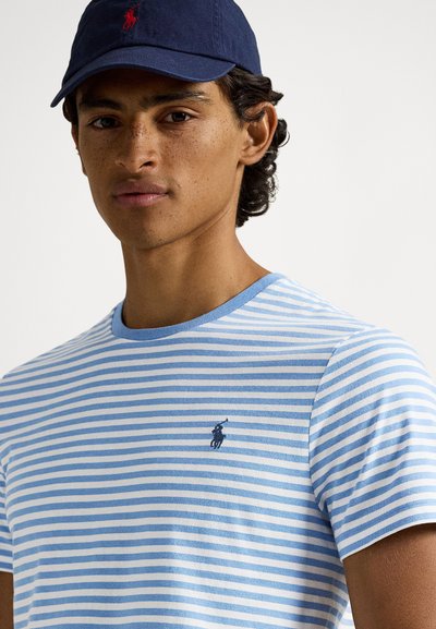 Ριγέ μπλουζάκι με στρογγυλή λαιμόκοψη σε ανοιχτό μπλε και λευκό βαμβάκι, με μπλε σκούρο λογότυπο στο στήθος. Συνδυάζεται με μπλε σκούρο καπέλο.