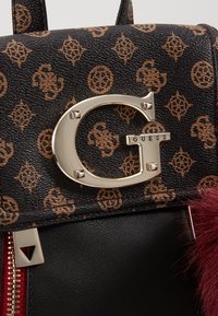 Sac à dos en similicuir à motif noir et marron avec un logo circulaire argenté bien visible, une fermeture éclair rouge et un accent en fourrure bordeaux.