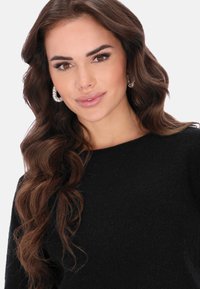 Fekete pulóver kerek nyakkal, texturált anyagból készült. A modellen hosszú, hullámos haj van, és kör alakú strasszköves fülbevalót visel.