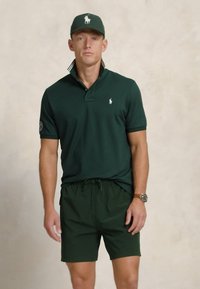 Polo shirt verde închis cu guler și logo pe piept, asortat cu șorturi verzi. Pălăria are un design de logo similar.