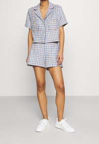 Persoon in een pastelgekleurde geruite korte blazer en bijpassende shorts met witte sneakers, staande op een lichtgrijze vloer en muur.