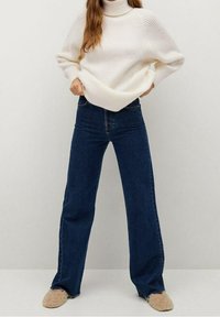 Kremowy prążkowany sweter z wysokim kołnierzem, zestawiony z szerokimi jeansami w ciemnoniebieskim kolorze. Modelka ma na sobie beżowe, pluszowe buty wsuwane.