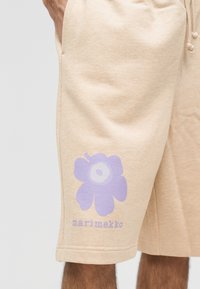 Beige shorts with lavender floral graphic and "marimekko" text on the left leg, worn by a person with tanned skin.