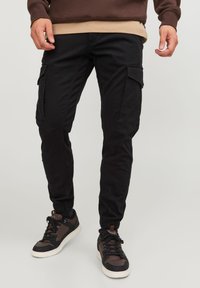 Svarta cargobyxor med elastiska muddar, flera fickor och en mjuk yta. Bärs med bruna och svarta sneakers.