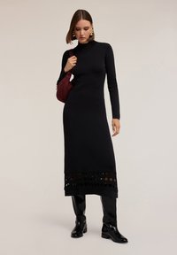 Donna in un lungo vestito nero a collo alto con orlo tagliato, stivali neri fino al ginocchio, orecchini dorati e una borsa a spalla bordeaux.