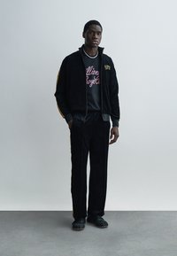 Μαύρο βελουτέ φόρμα με κίτρινες ρίγες. Το μπουφάν έχει φερμουάρ και τυπωμένο λογότυπο. Το μοντέλο φοράει μαύρο γραφικό t-shirt και μαύρα αθλητικά παπούτσια.