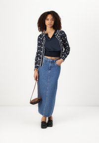 Navy patterned cardigan with white accents, layered over a dark blue top. Long denim skirt in light blue, paired with black loafers and a brown bag.
