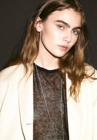 Ein Modell trägt ein durchscheinendes schwarzes Oberteil, das unter einer hellen cremefarbenen Jacke geschichtet ist. Langes braunes Haar und eine zarte silberne Halskette runden den Look ab.