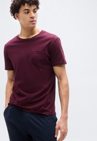 Νέος άνδρας με σγουρά μαλλιά, φορώντας μπορντό κοντομάνικη μπλούζα με τσέπη στο στήθος και σκούρο μπλε παντελόνι, κοιτάζει προς τα αριστερά του.