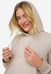 Haut à capuche beige clair avec une coupe décontractée, doté d'une capuche avec cordon de serrage et d'accents en cordon texturé. Tissu doux et lisse avec des poignets.