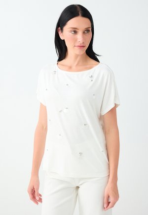Femme aux cheveux noirs portant un haut blanc à manches courtes avec de petites décorations florales et un pantalon blanc devant un fond clair uni.