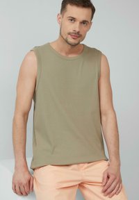 Ärmelloses olivgrünfarbenes Tanktop aus geripptem Stoff, mit Rundhalsausschnitt und lässiger Passform, kombiniert mit hellpeachfarbenen Hosen.