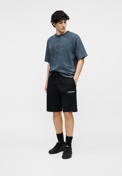 Jeune homme portant un t-shirt bleu délavé "Off-White", un short noir à cordon avec texte blanc, des chaussettes noires et des baskets noires, debout les mains dans les poches.