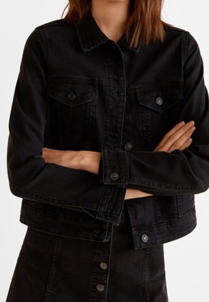 Person trägt eine schwarze Jeansjacke mit geknöpften Brusttaschen und verschränkten Armen vor dem Oberkörper.