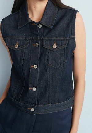 Ærmeløs mørkeblå denimvest med sølvknapper og brystlommer med klap, båret over mørke bukser mod en lys baggrund.