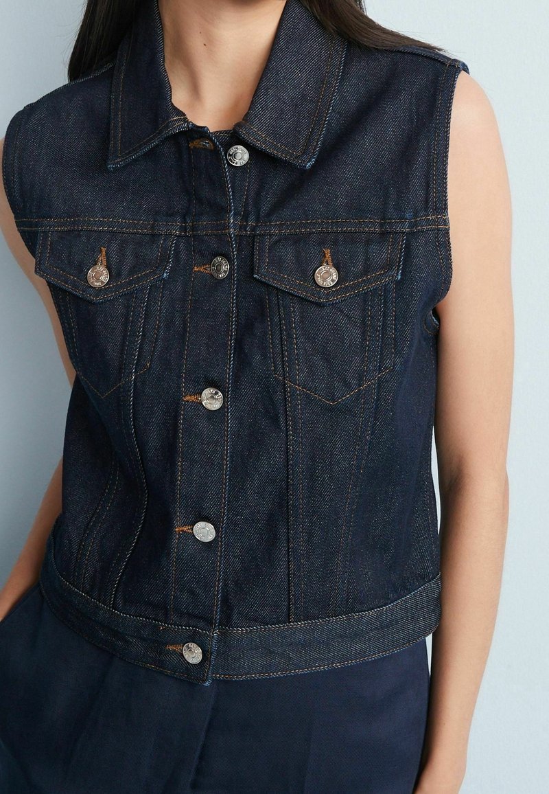 Veste en jean bleu foncé sans manches avec des boutons argentés et des poches à rabat sur la poitrine, portée sur un pantalon foncé sur un fond clair.