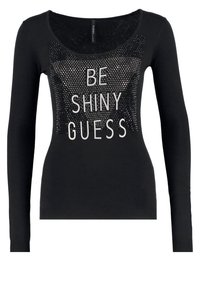 Chemise noire à manches longues avec encolure ronde, ornée de petits clous brillants et d'un texte blanc indiquant "BE SHINY GUESS" sur le devant.