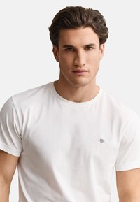 Young man with short dark hair wearing a plain white crew neck t-shirt with a small emblem on the left chest against a neutral background.