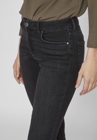 Pantalones vaqueros negros de mezclilla con un diseño ajustado, estilo de cinco bolsillos y detalles en tono plateado en la cintura y los bolsillos, mostrados a la altura de la cintura.