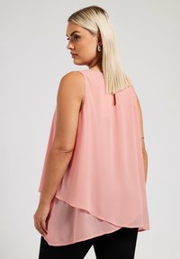 Ljusrosa chiffontopp utan ärmar med en nyckelhålsdetalj på ryggen och en asymmetrisk fåll, kombinerad med svarta byxor. Slen yta.