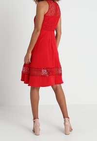 Robe rouge avec des détails en dentelle au niveau du corsage et de l'ourlet, accompagnée d'une jupe fluide. Pour finir le look, des chaussures à talons hauts nudes.