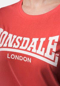 Red cotton t-shirt featuring a white text graphic "LONSDALE LONDON." Crew neck design with a smooth texture and short sleeves.